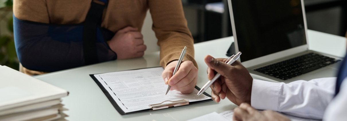 Close up of man filling in medical insurance form, injured hand in sling in background, copy space