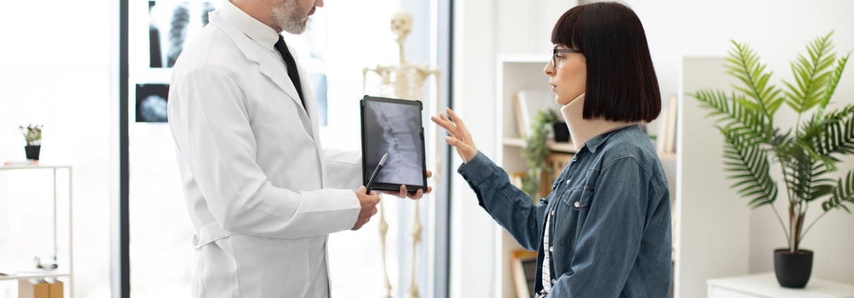 Confident mature man in lab coat holding digital tablet while young lady in cervical collar sitting in exam room. General practitioner showing test results to female with car accident injury.