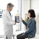 Confident mature man in lab coat holding digital tablet while young lady in cervical collar sitting in exam room. General practitioner showing test results to female with car accident injury.